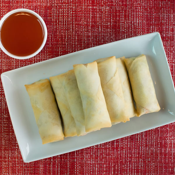 Six crispy Dragon Dynasty egg rolls on a white plate with a side of sweet and sour sauce.